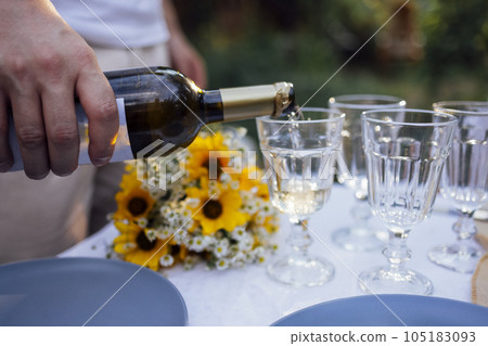 Man pours white wine from dark bottle into glasses outdoors in garden. Closeup of male hand and goblet. 105183093