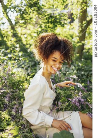 Attractive mixed race girl in white elegant chiffon dress sits among blooming purple flowers. 105183094