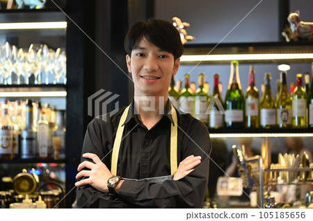 Portrait of male bartender wearing apron standing with arm crossed at the bar counter in restaurant and smiling at camera Portrait of male bartender wearing apron standing with arm crossed at the bar counter in restaurant and smiling at camera 105185656