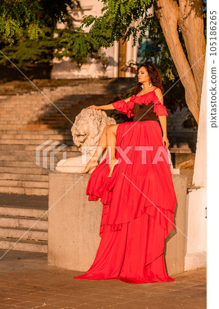A woman in a long red dress against the backdrop of sunrise, bright golden light of the sun's rays. The concept of femininity, harmony. 105186265