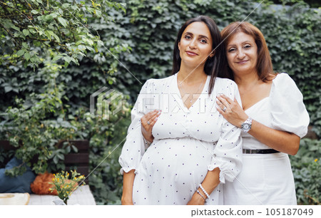 Attractive pregnant brunette has her mom cuddling outdoors in the garden. 105187049