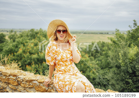 Attractive girl in pink dress and wide brimmed straw hat smiles and enjoys nature against backdrop of beautiful summer landscape 105189199