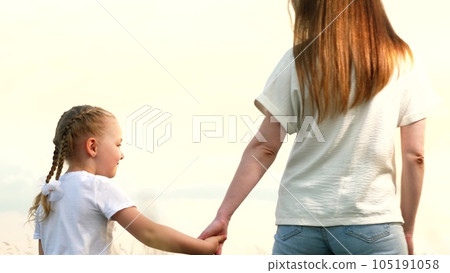 Mother and little daughter walk joining hands across field at sunset light backside view. Happy daughter holds mother hand walking together on country meadow. Mother and daughter enjoy family time 105191058