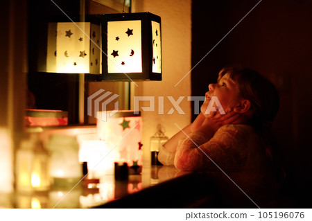 Little kid girl sitting by window with selfmade hand crafted lanterns with candles for St. Martin procession. Toddler child looking at glowing lantern. German tradition Martinsumzug. Home decoration 105196076