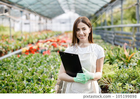 Portrait of gardener woman at work in greenhouse with notebook examines the growing flowers in greenhouse. Home gardening, love of plants and care. Small business. Portrait of gardener woman at work in greenhouse with notebook examines the growing flowers in greenhouse. Home gardening, love of plants and care. Small business. 105199898