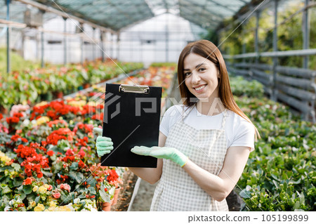Portrait of gardener woman at work in greenhouse with notebook examines the growing flowers in greenhouse. Home gardening, love of plants and care. Small business. 105199899