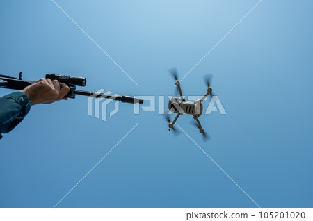 A man aims to shoot a rifle at a flying drone against a blue sky. A man aims to shoot a rifle at a flying drone against a blue sky. 105201020