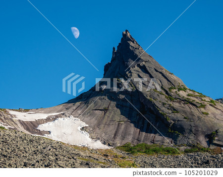 Pointed cliff with a small snow glacier, moon in the blue evening sky. Awesome scenic mountain landscape with big pointed stones in evening. Sharp rocks background. Western Sayans. 105201029