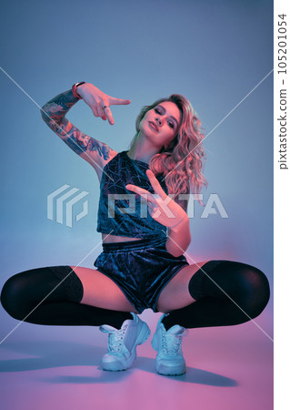 Young fitness blonde woman wearing blue velour booty shorts squats in front of camera, looking at camera, pink and blue background Young fitness blonde woman wearing blue velour booty shorts squats in front of camera, looking at camera, pink and blue background 105201054