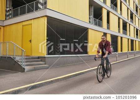 New bike. Handsome man with stubble in casual clothes riding on his bicycle along a road against yellow building 105201096