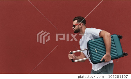 Ready for a journey. Side view of handsome bearded man in sunglasses holding his luggage while standing against red wall outdoors Ready for a journey. Side view of handsome bearded man in sunglasses holding his luggage while standing against red wall outdoors 105201107