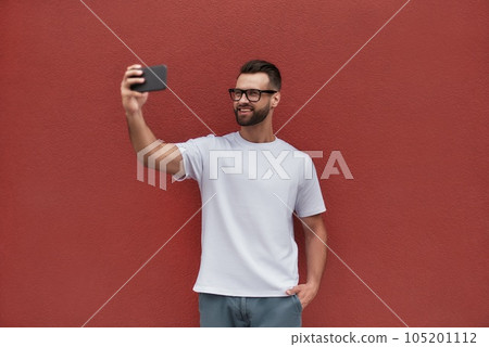 Happy moments. Handsome young man in casual clothes making selfie with his mobile phone and smiling while standing against red wall outdoors Happy moments. Handsome young man in casual clothes making selfie with his mobile phone and smiling while standing against red wall outdoors 105201112