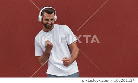 Good mood Portrait of happy handsome man in headphones listening to the music and dancing against red wall outdoors 105201124