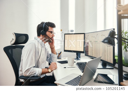 Always available. Side view of young bearded trader in headset talking with client and looking at monitor screen with trading charts and financial data while sitting in his modern office. Always available. Side view of young bearded trader in headset talking with client and looking at monitor screen with trading charts and financial data while sitting in his modern office. 105201145
