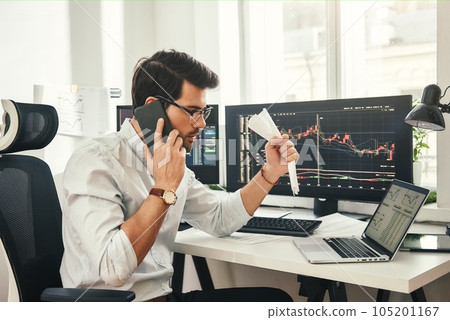 Stock falling. Shocked bearded businessman or trader in formalwear and eyeglasses is talking with client by smartphone and feeling nervous while sitting in modern office. 105201167