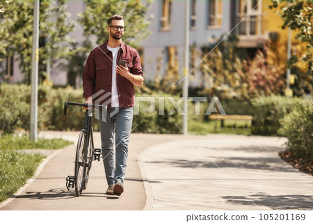 My way. Handsome man with stubble in casual clothes and eyeglasses pulling his bicycle and holding smartphone while walking outdoors 105201169