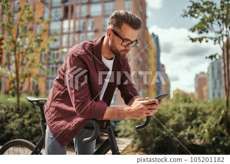 Surfing the net. Side view of handsome man with stubble in casual clothes and eyeglasses leaning at his bicycle and looking at mobile phone while standing outdoors Surfing the net. Side view of handsome man with stubble in casual clothes and eyeglasses leaning at his bicycle and looking at mobile phone while standing outdoors 105201182