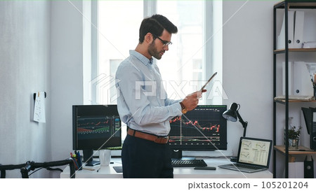 Analyzing data. Successful young trader in formal wear is looking at trading graphs and reports on his digital tablet while standing in front of computer screens with trading charts in the office. Analyzing data. Successful young trader in formal wear is looking at trading graphs and reports on his digital tablet while standing in front of computer screens with trading charts in the office. 105201204