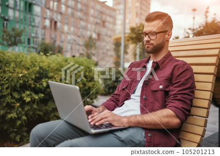 Modern blogger. Side view of young handsome man with stubble in casual clothes and eyeglasses working on laptop while sitting on the bench outdoors 105201213