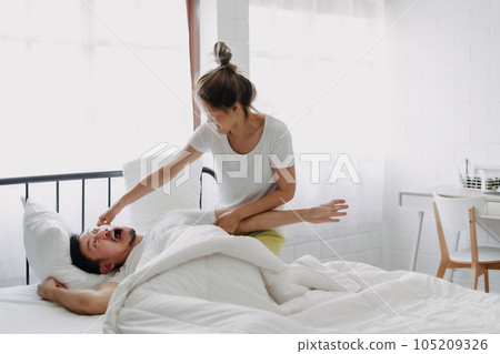 Funny couple wife try to wake lazy husband up in white bedroom. Funny couple wife try to wake lazy husband up in white bedroom. 105209326