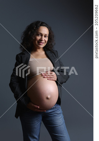 Portrait of a pregnant woman in gray blazer and beige bra, stroking her tummy, smiling and looking confidently at camera 105209662