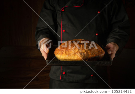 The cook is holding fresh rye wheat loaf of bread on a sheet pan. Copy space. The concept of baking bread at home The cook is holding fresh rye wheat loaf of bread on a sheet pan. Copy space. The concept of baking bread at home 105214451