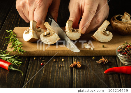 The cook cuts Agaricus mushrooms on a cutting board. Close-up of a chef hands with a knife. Dark space for recipe or menu 105214459