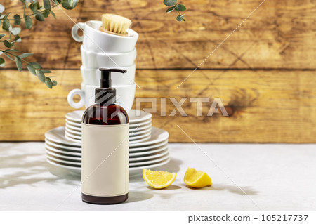 Mockup of dish detergent. Eco friendly natural cleaning dishes. Bottle with dish detergent with clean plates and cups on wooden background with sprig of eucalyptus. Front view, copy space 105217737