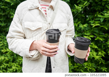 A girl in a residential neighborhood holds cups of coffee. Two glasses of hot drinks in the hands of a young woman. Breakfast on the go. Picnic and going out concept.  105221095