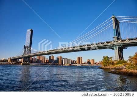 View of entire Manhattan Bridge from Brooklyn in New York City over waters with blue sky 105230999