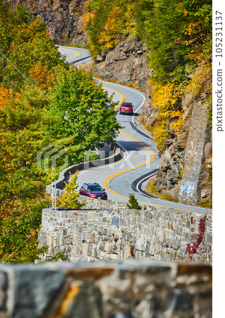 Winding road with cars and stone wall going up through cliffs and forest 105231137
