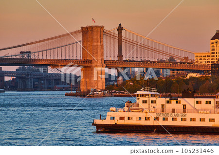 Governors Island Ferry in golden hour sunset dusk light from waters passing by Brooklyn Bridge with American Flag 105231446