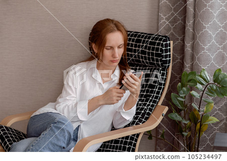 A young happy smiling pretty woman sitting in an armchair holding a smartphone, using modern cellular technology, looking at her cell phone while working remotely. High quality photo A young happy smiling pretty woman sitting in an armchair holding a smartphone, using modern cellular technology, looking at her cell phone while working remotely. High quality photo 105234497