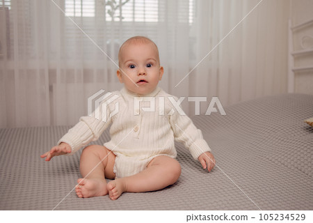 A cute baby in a white knit bodysuit sits on the bed. High quality photo A cute baby in a white knit bodysuit sits on the bed. High quality photo 105234529