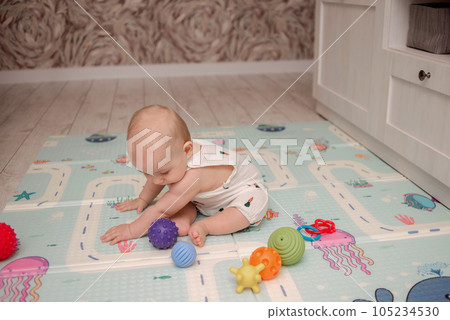 A cute baby in a white suit sits on a soft mat and plays with bright toys. High quality photo 105234530