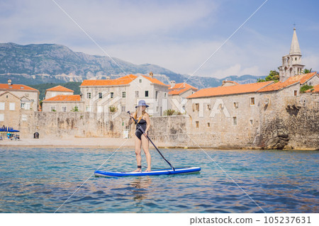 Young women Having Fun Stand Up Paddling in blue water sea in Montenegro. Against the backdrop of the Old Town of Budva. SUP 105237631