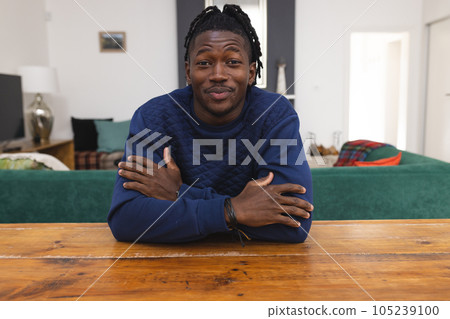 African american man with arms crossed talking over video call while sitting at table in living room 105239100