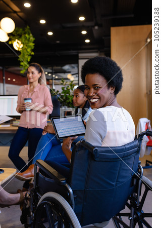 Portrait of happy african american casual businesswomen in wheelchair holding tablet in office 105239188