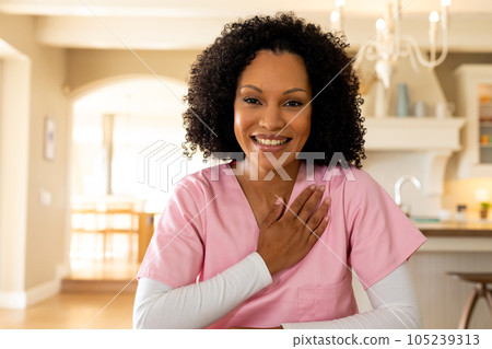 Happy biracial female medical worker wearing scrubs having video call in kitchen at home, listening 105239313