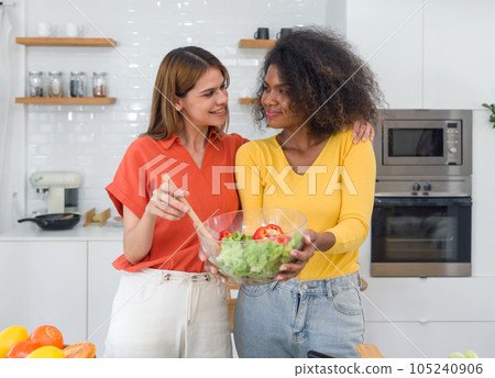 Two women standing in a kitchen, holding a bowl of salad. The kitchen is well-lit, with a countertop, cabinets, and a oven visible in the background. Two women standing in a kitchen, holding a bowl of salad. The kitchen is well-lit, with a countertop, cabinets, and a oven visible in the background. 105240906