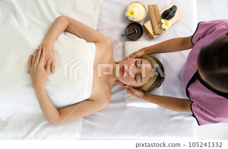 A serene spa scene featuring a relaxed woman lying on a massage table, a trained professional applying soothing technique to her face. 105241332