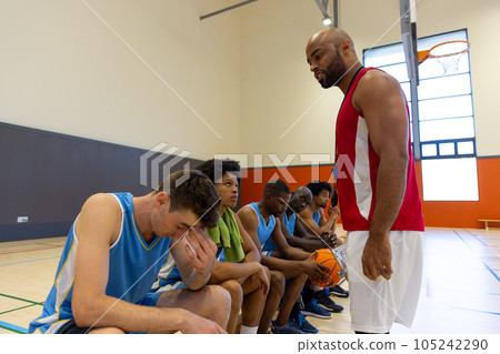 Diverse male basketball players discussing game plan with coach, sitting on bench at gym 105242290