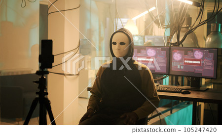 Male spy with anonoymous mask filming live threat video, asking for ransomware instead of leaking important data. Young person threatening to expose information, online cybercrime. 105247420