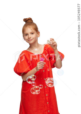 Portrait of little redhead girl, child in red dress blowing soap bubbles against white studio background 105249377
