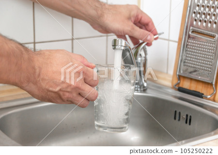 Filling a glass with tap water for drinking by holding it in man hand at the sink in the kitchen Filling a glass with tap water for drinking by holding it in man hand at the sink in the kitchen 105250222