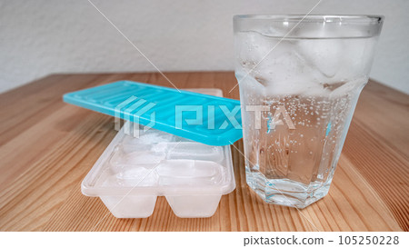 Sparkling water with bubbles in a big transparent glass filled with ice cubes and a soft plastic blue form to make ice cubes in the freezer. Solution to overcome extreme high temperature in summer 105250228