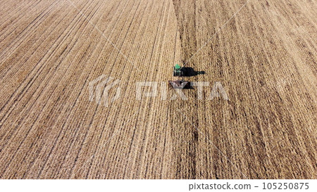Tractor plowing the ground. Flying over green tractor that plows up ground in yellow field after harvesting wheat on autumn sunny day. Tractor digging land. Agricultural field work. Aerial drone view Tractor plowing the ground. Flying over green tractor that plows up ground in yellow field after harvesting wheat on autumn sunny day. Tractor digging land. Agricultural field work. Aerial drone view 105250875