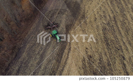 Tractor plowing the ground. Flying over green tractor that plows up ground in yellow field after harvesting wheat on autumn day. Tractor digging land. Agricultural work on field. Aerial drone view Tractor plowing the ground. Flying over green tractor that plows up ground in yellow field after harvesting wheat on autumn day. Tractor digging land. Agricultural work on field. Aerial drone view 105250879