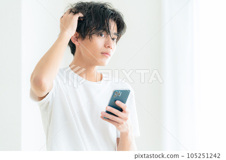 一位年輕人在客廳窗邊一邊操作智能手機一邊護理頭髮 一位年輕人在客廳窗邊一邊操作智能手機一邊護理頭髮 105251242