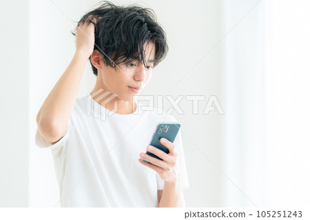 一位年輕人在客廳窗邊一邊操作智能手機一邊護理頭髮 一位年輕人在客廳窗邊一邊操作智能手機一邊護理頭髮 105251243